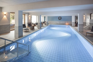 A large indoor swimming pool with a tiled floor, surrounded by lounge chairs and a staircase leading to an upper level. - Landhotel Saarschleife (Mettlach)