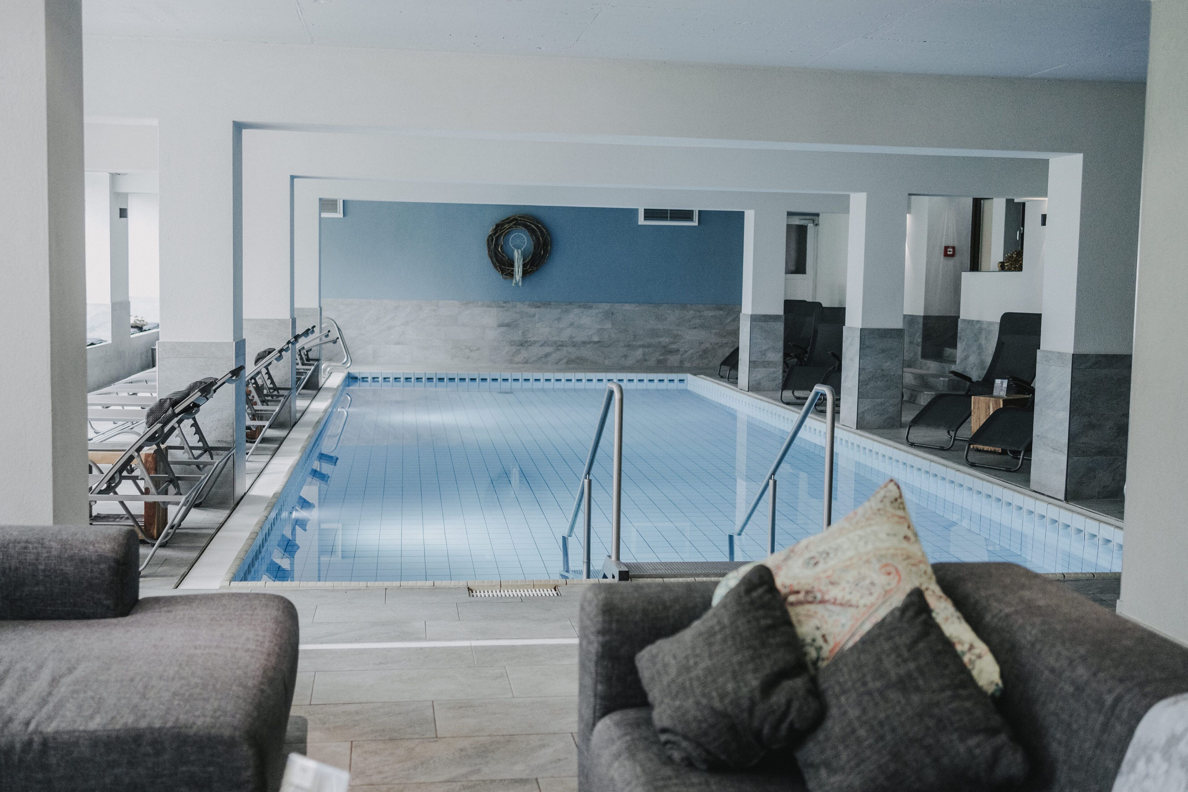 A modern indoor swimming pool with a clear view of the pool area, seating arrangements, and a water feature on the wall.