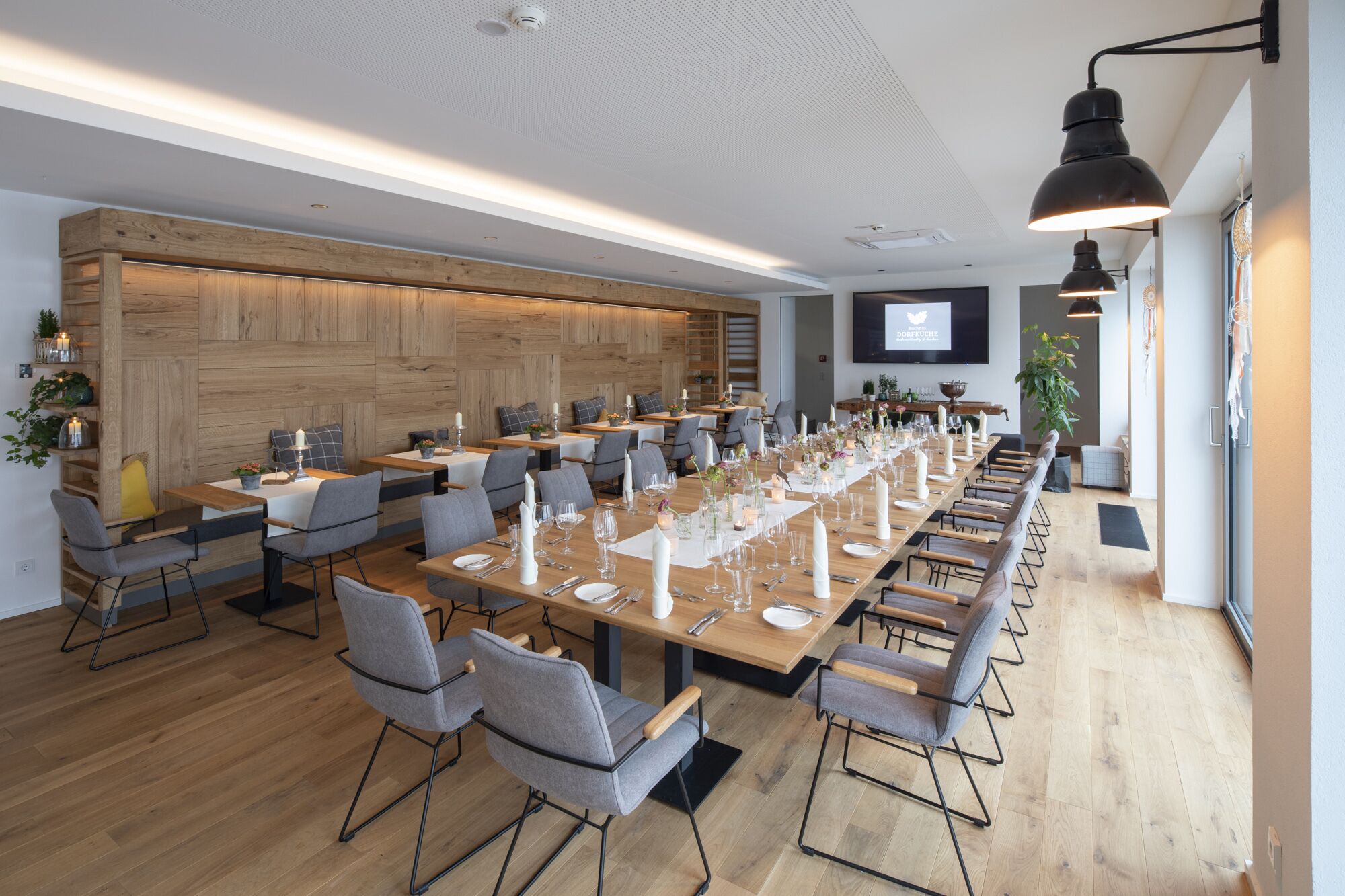 A conference room with long tables, grey chairs, wooden panel walls, and a mounted TV screen.
