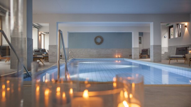 A modern indoor swimming pool with a clear view of the water, a glass railing, and a few lit candles on the poolside. - Landhotel Saarschleife (Mettlach)