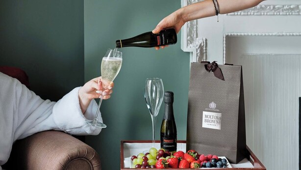 A person pouring champagne into a glass, a bottle of Prosecco, and a box of assorted fruits on a table. - Montagu Place Hotel (London)
