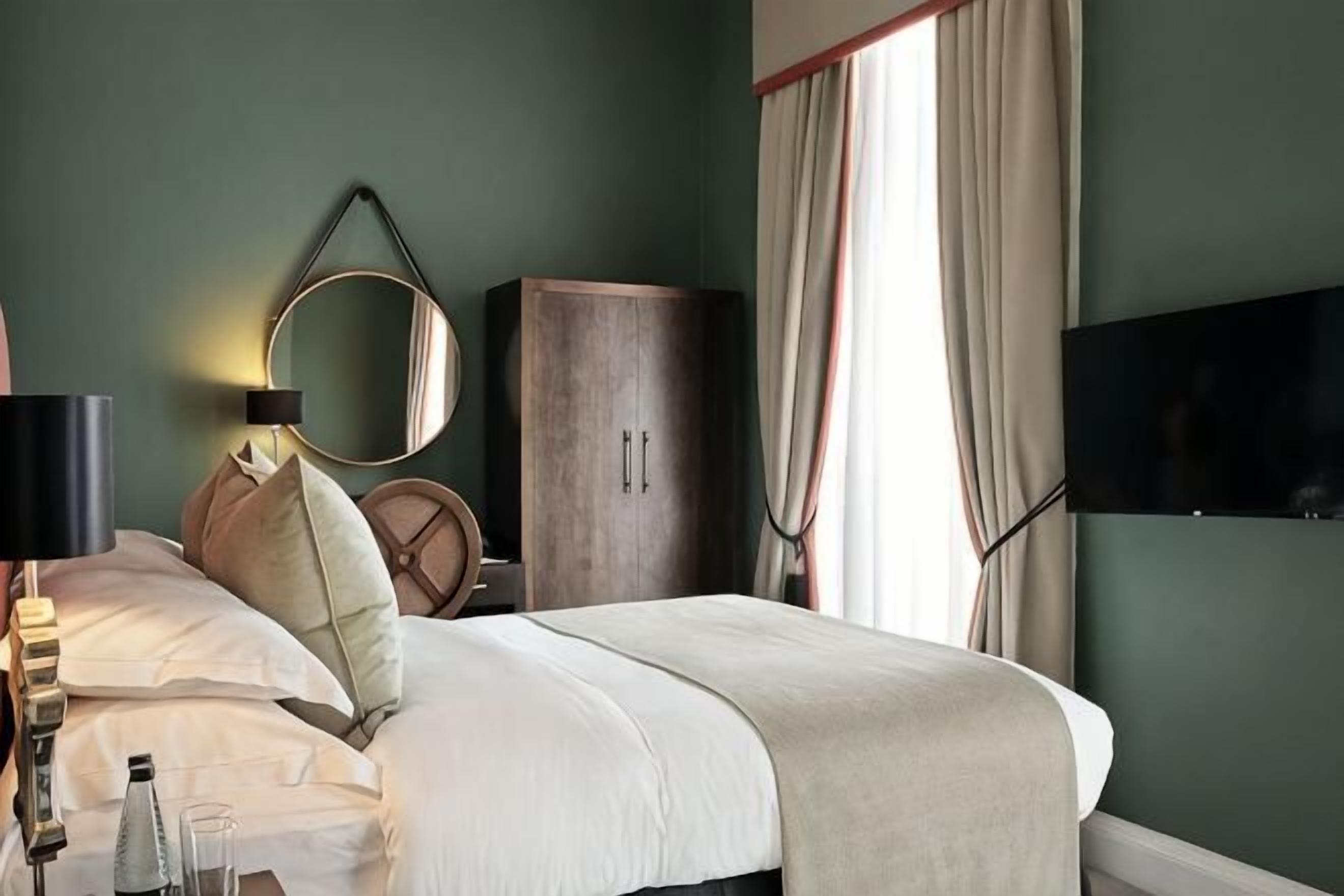 A neatly made bed with pillows, a bedside lamp, a mirror, a wardrobe, and a television mounted on the wall.