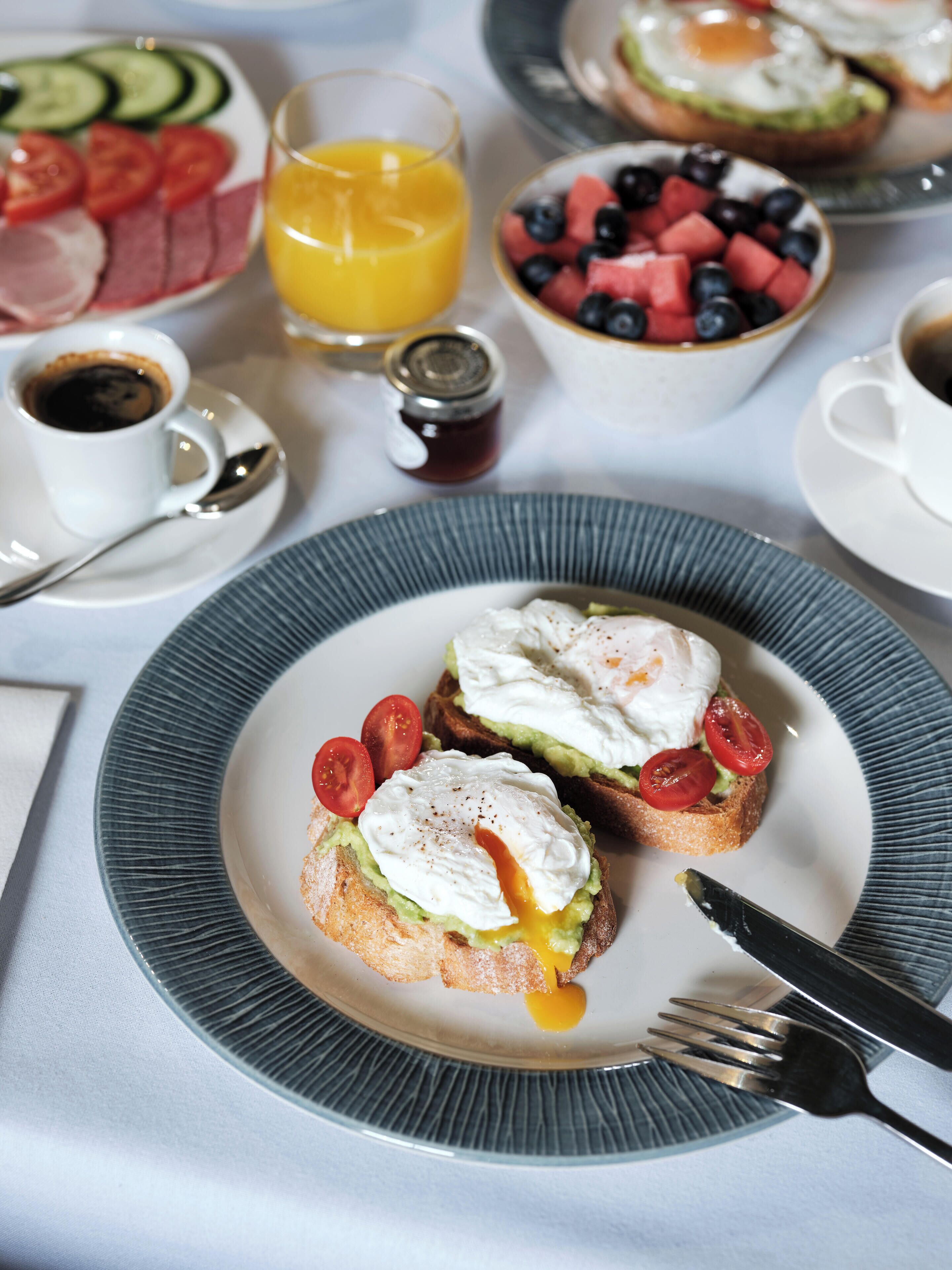 Een ontbijttafel met toast belegd met eieren en tomaten, een fruitschaal, een glas sinaasappelsap en een kop koffie.