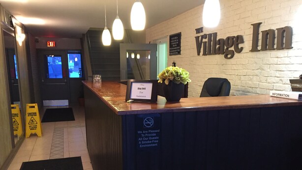 Un espace d’accueil avec un comptoir en bois, une enseigne indiquant « The Village Inn » et une autre indiquant « Sonnez pour obtenir de l’aide ».