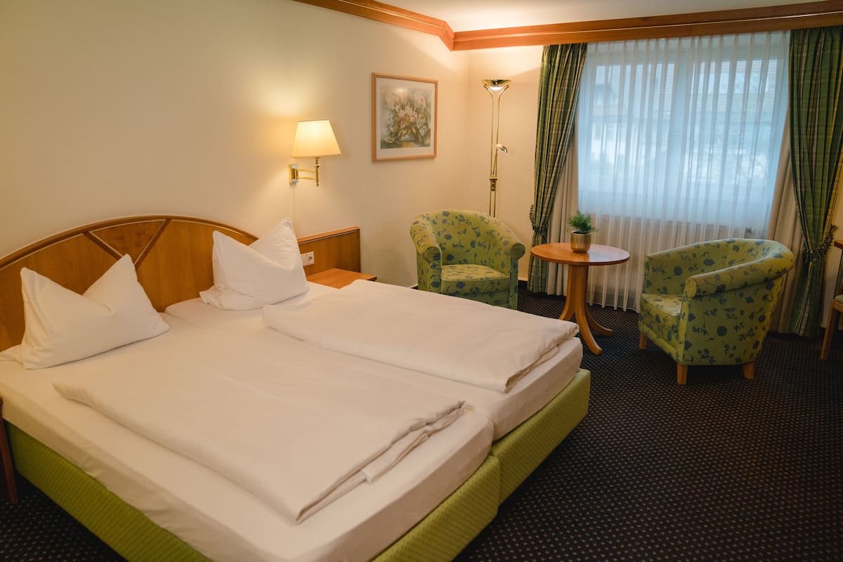 A hotel room with a large bed, two armchairs, a small table, a lamp, and a window with curtains.