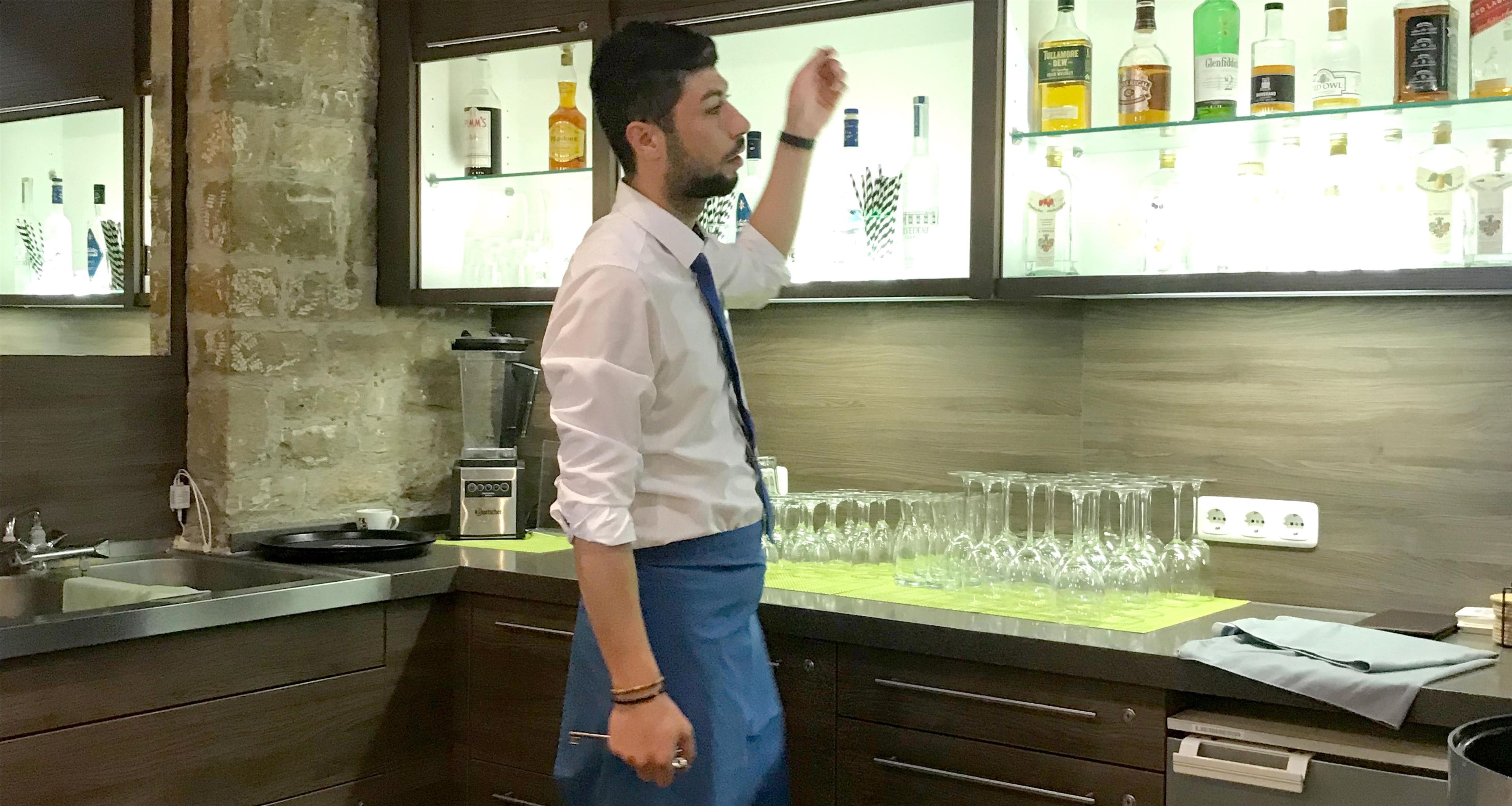 A person standing in a kitchen with a countertop displaying various bottles, a blender, and a sink.