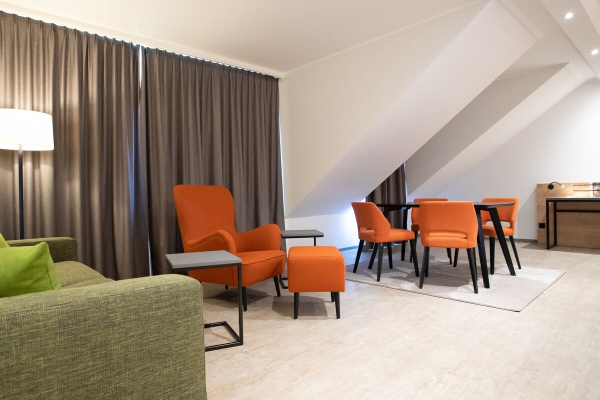 A modern hotel room with a green sofa, orange chairs, a small table, and a desk with a lamp.