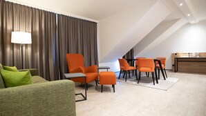 A modern hotel room with a green sofa, orange chairs, a small table, and a desk with a lamp.