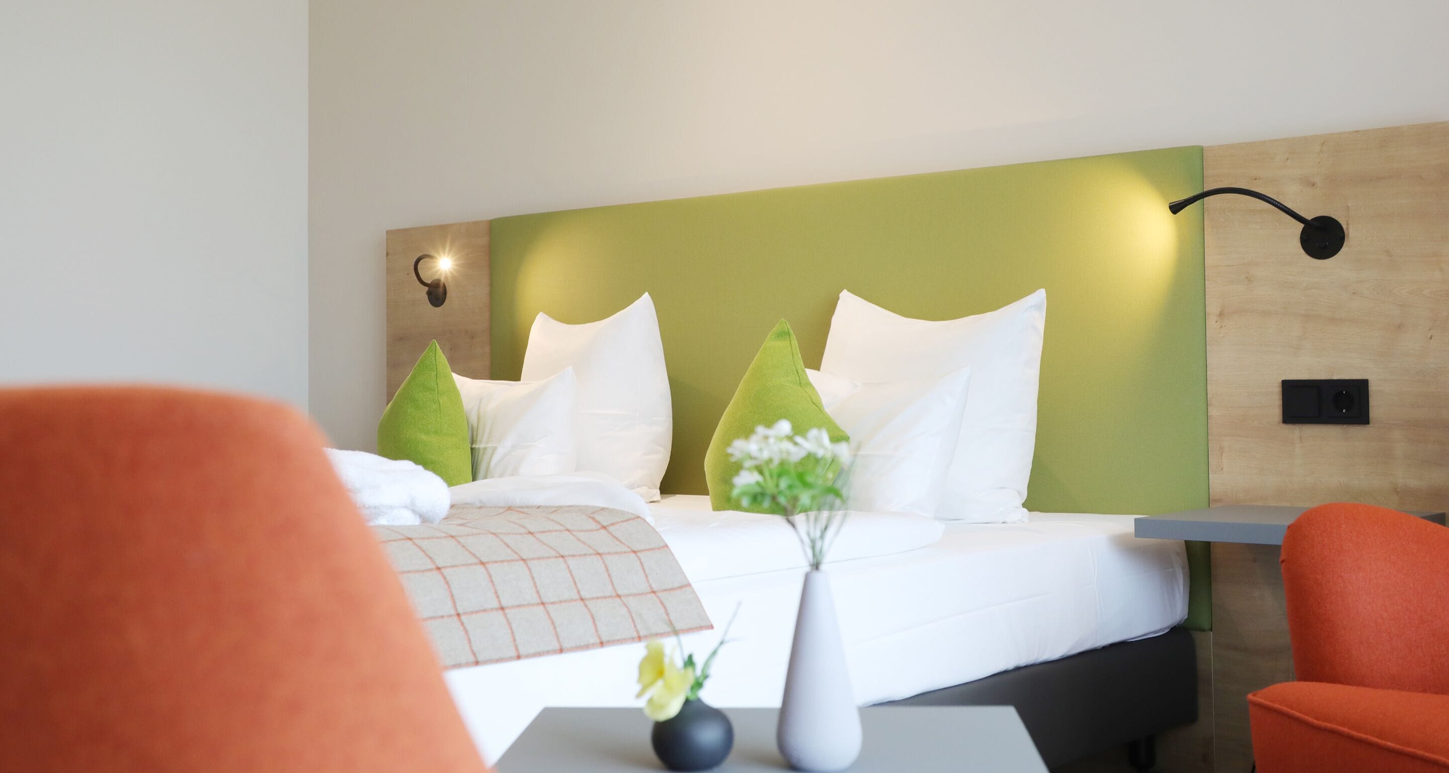 Une chambre d’hôtel avec un lit, un mur vert, des oreillers blancs, un vase avec des fleurs et des chaises orange.