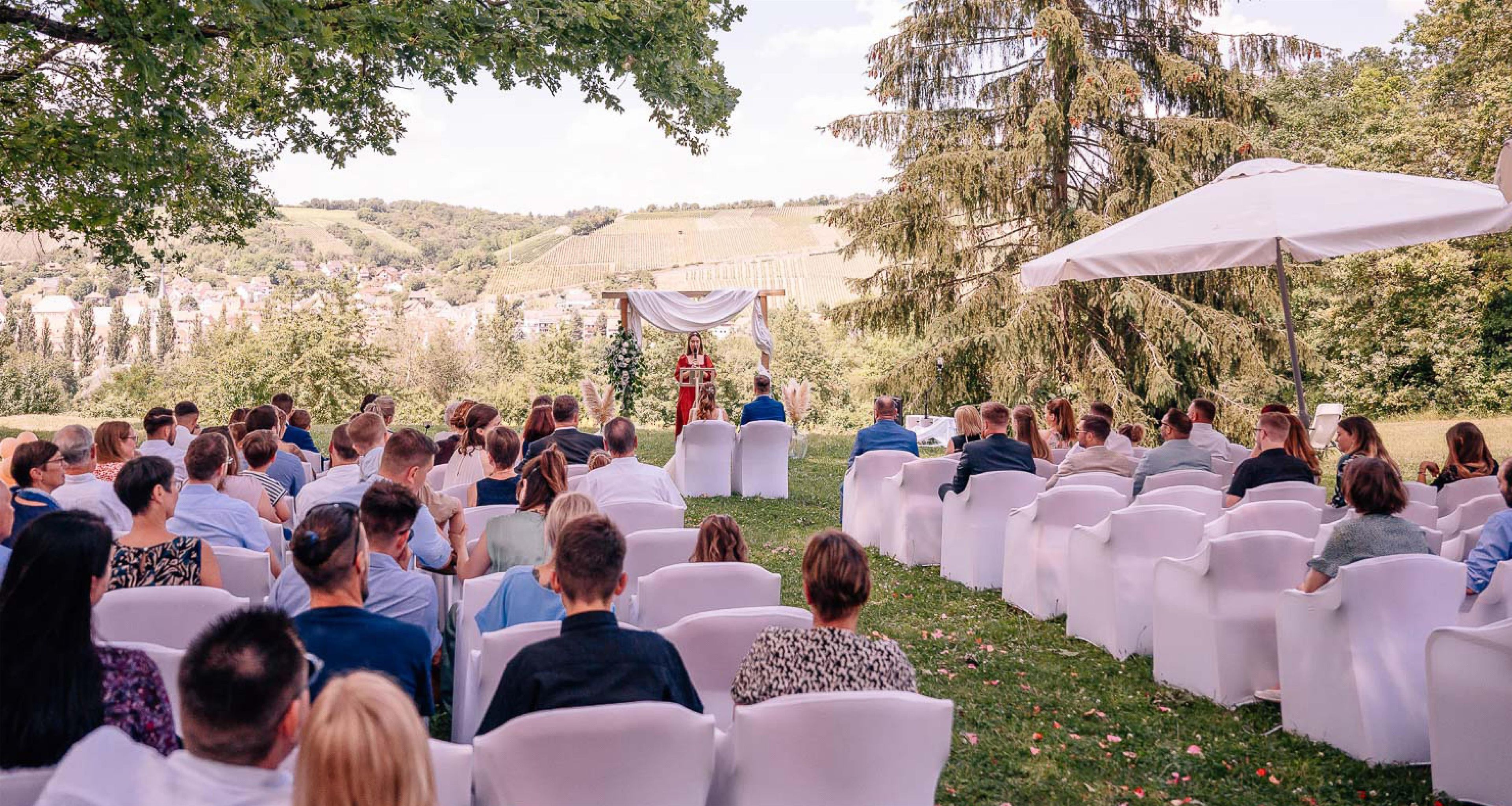 An outdoor wedding ceremony with guests seated on white chairs facing a speaker.