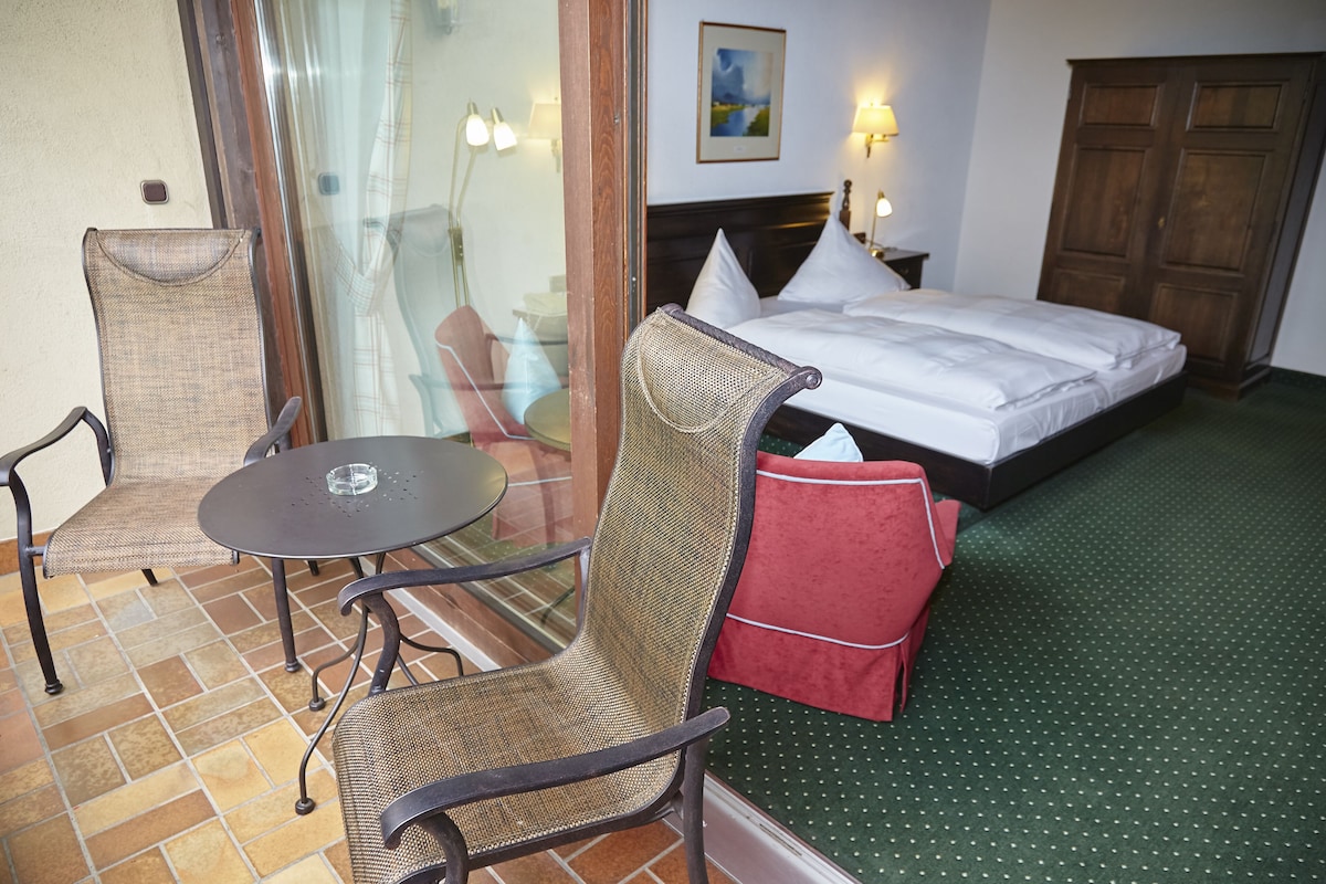 A hotel room with a bed, a chair, a small table, and a view of the outdoors through a glass door.