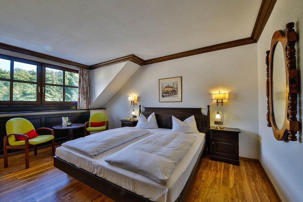 A bedroom with a large bed, two bedside tables with lamps, a chair, a small table, and a window with a view of greenery.