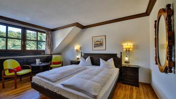 A bedroom with a large bed, two bedside tables with lamps, a chair, a small table, and a window with a view of greenery.