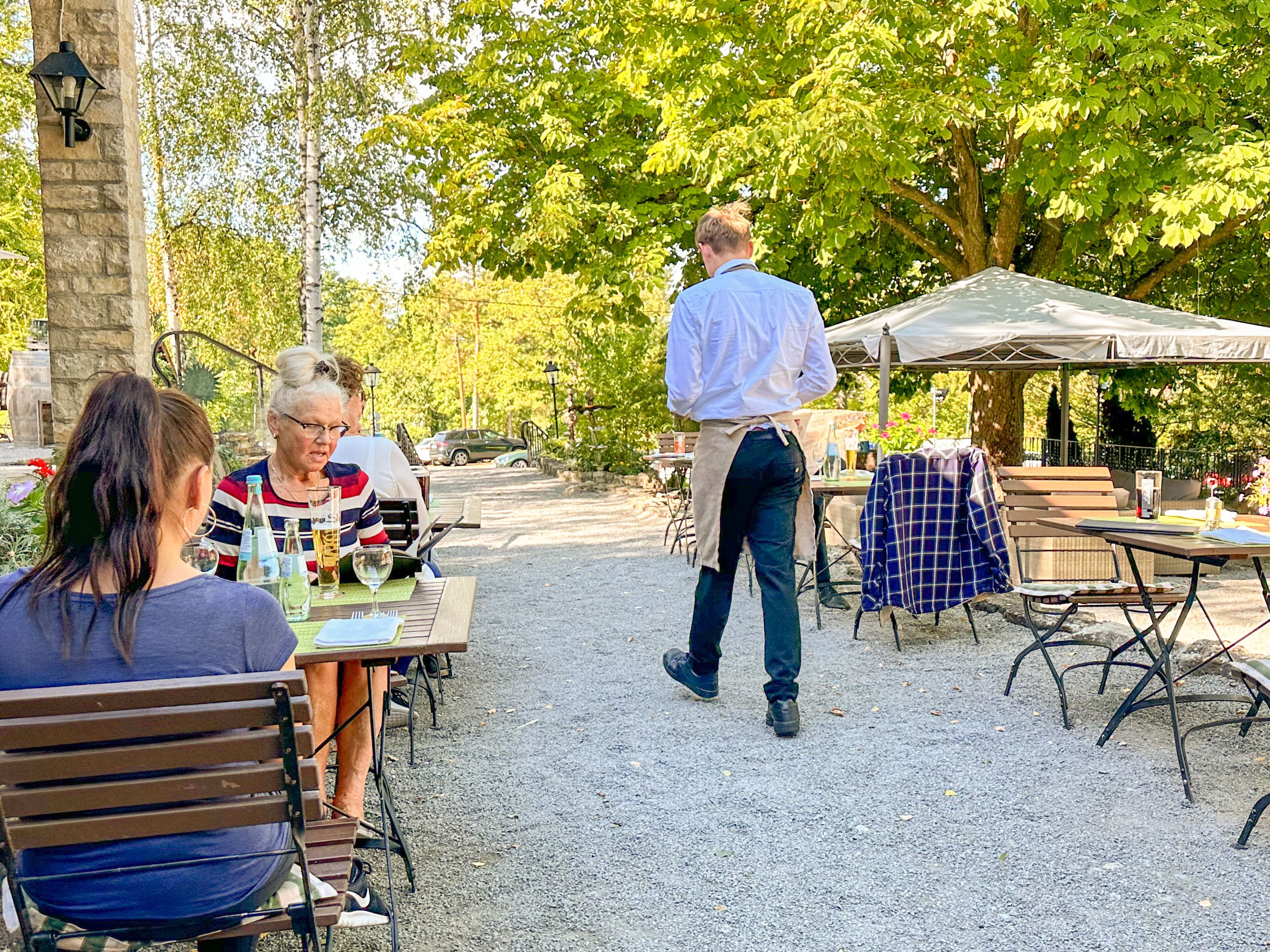 An outdoor café with tables and chairs, surrounded by trees and a gravel path.
