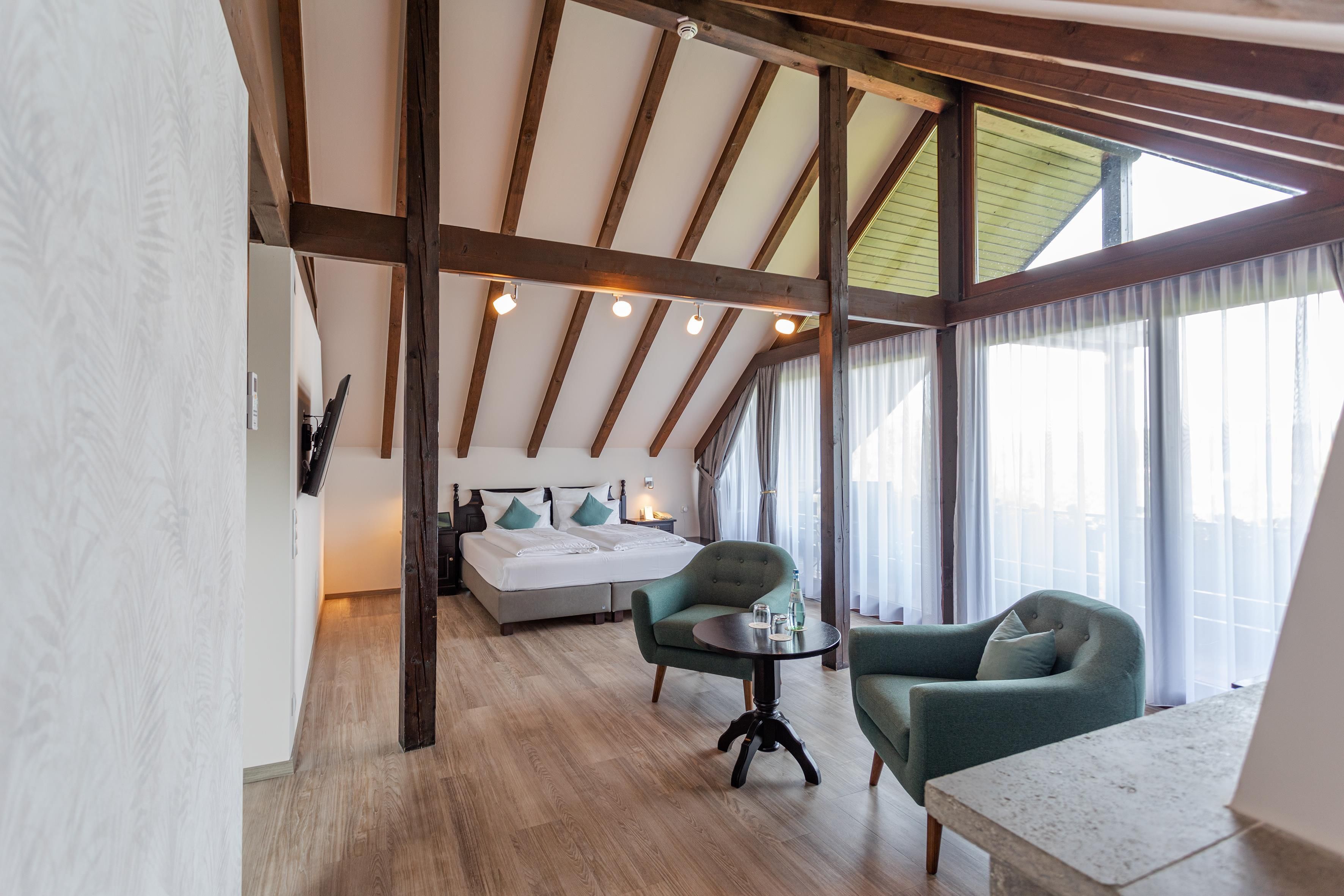 Habitación de hotel con techo estilo loft, una cama con dos almohadas, un sillón verde, una mesita pequeña y un ventanal grande con cortinas.
