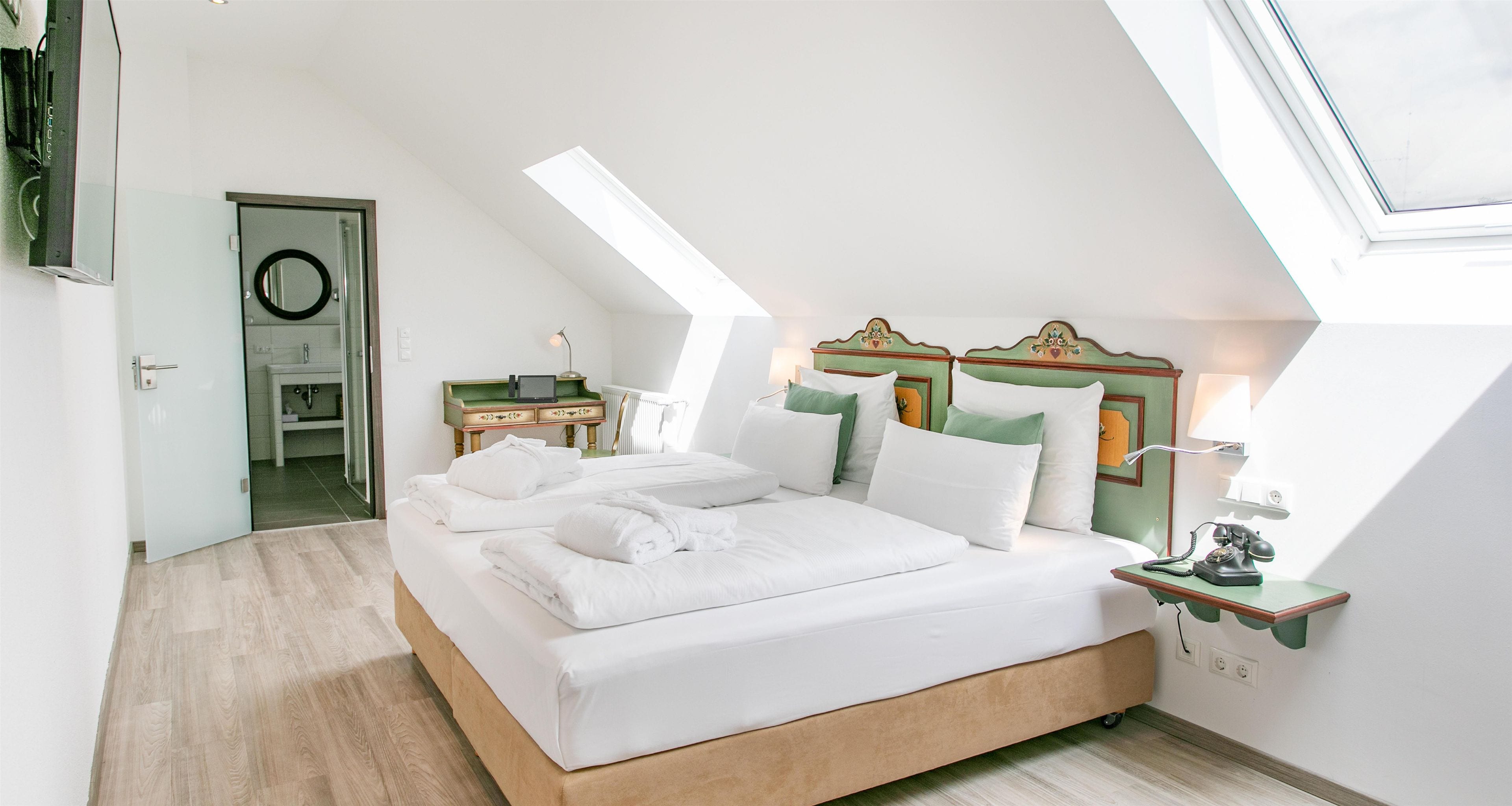 Ein Schlafzimmer mit schrägem Dach, einem Bett mit weißer Bettwäsche, einem Nachttisch mit Lampe und einem kleinen Schreibtisch mit Computer.