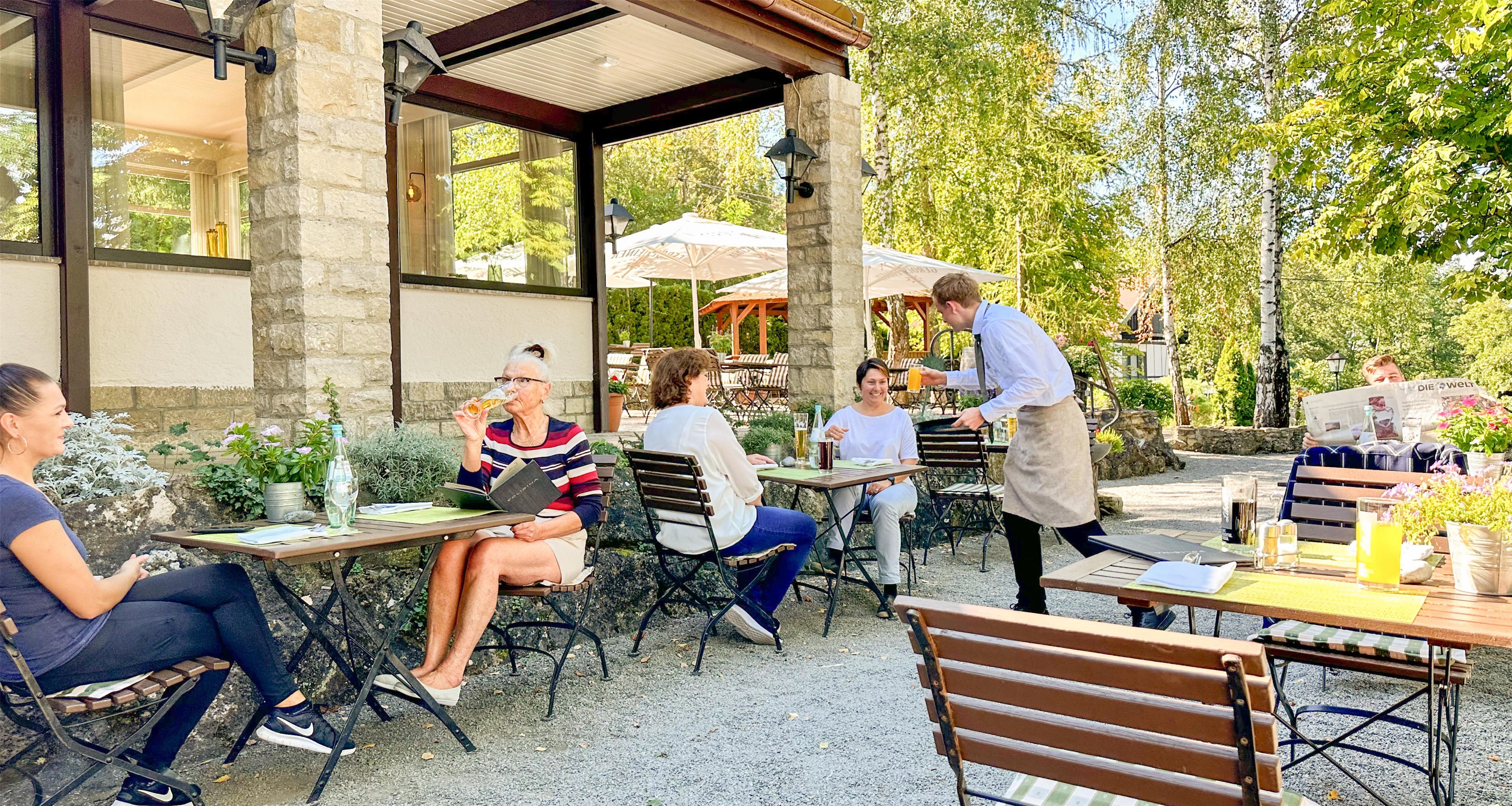 An outdoor café with tables and chairs, a waiter attending to guests, and a stone building in the background.