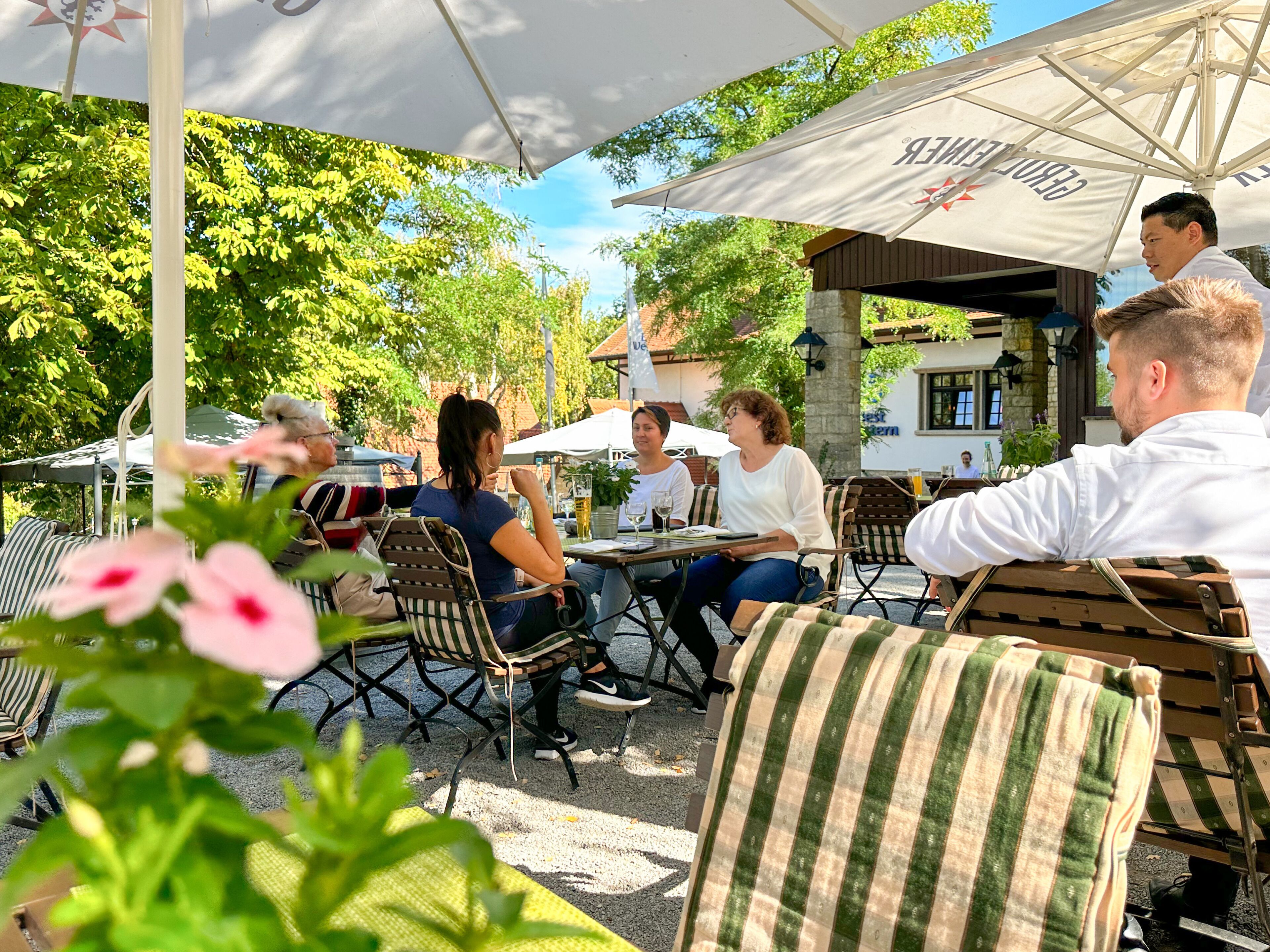 An outdoor café with tables and chairs under umbrellas, surrounded by greenery.