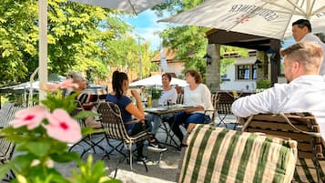 An outdoor café with tables and chairs under umbrellas, surrounded by greenery.