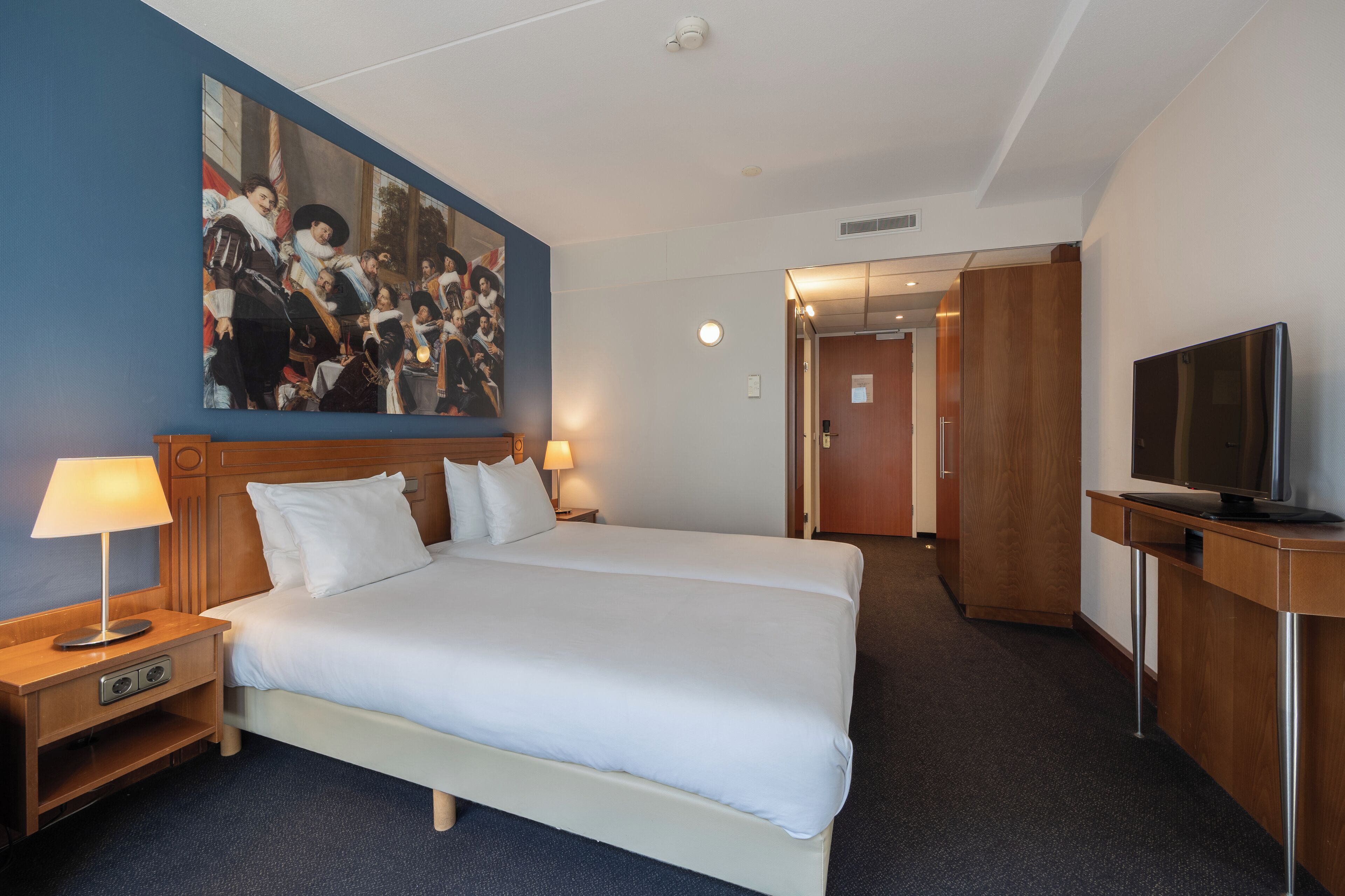 A hotel room with a large bed, a desk with a TV, a painting on the wall, and a door leading to another room.