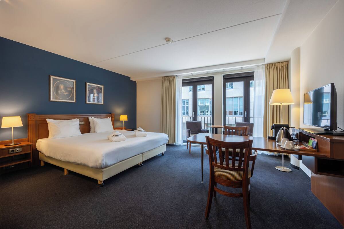 A hotel room with a bed, a desk, a chair, a TV, and two windows with curtains.