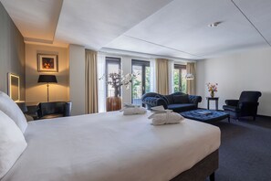 Ein Hotelzimmer mit einem großen Bett, einem Sofa, einem Sessel und einem kleinen Tisch. An den Fenstern hängen Vorhänge, und auf dem Nachttisch steht eine Lampe.