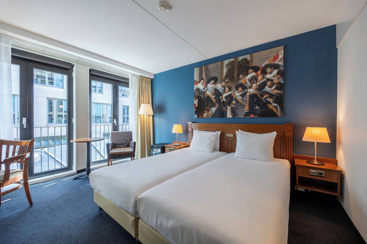 A hotel room with two beds, a large window, a balcony, and a painting on the wall.