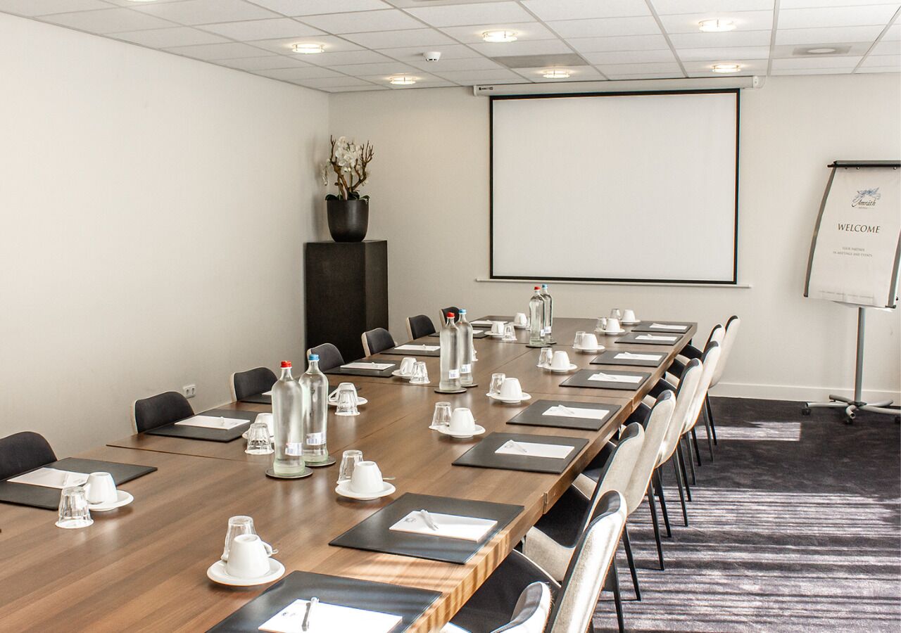Een vergaderzaal met een lange tafel, stoelen, waterflesjes en een projectiescherm.