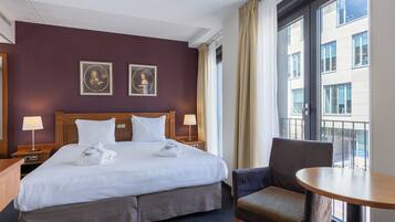 Quarto de hotel com uma cama grande, duas mesas de cabeceira com luminárias, uma cadeira e uma varanda com vista para edifícios.