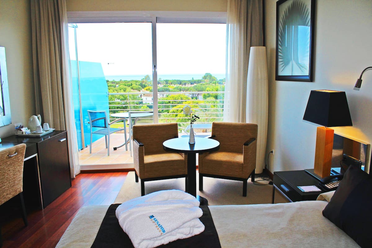 A hotel room with a bed, a desk, a chair, a small table, a lamp, and a view of the outdoors.
