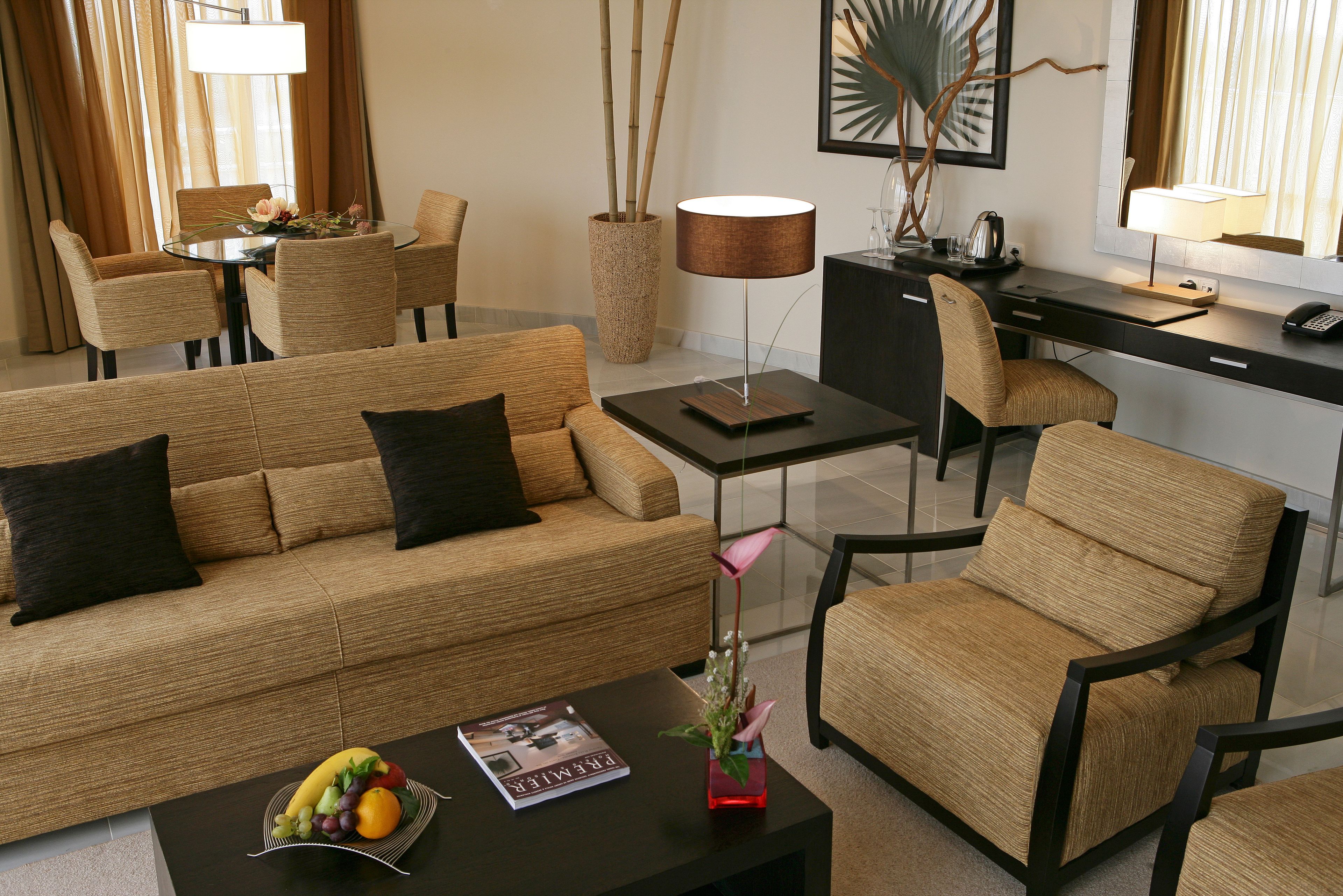 A modern hotel room with a sofa, chairs, a dining table, a desk, and a coffee table with a magazine and fruit.