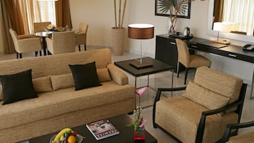 Una habitación de hotel moderna con sofá, sillas, una mesa de comedor, un escritorio y una mesa de centro con una revista y fruta.