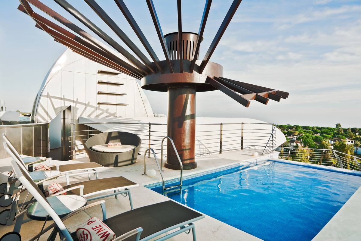 A rooftop pool area with lounge chairs, a hot tub, and a view of the city.