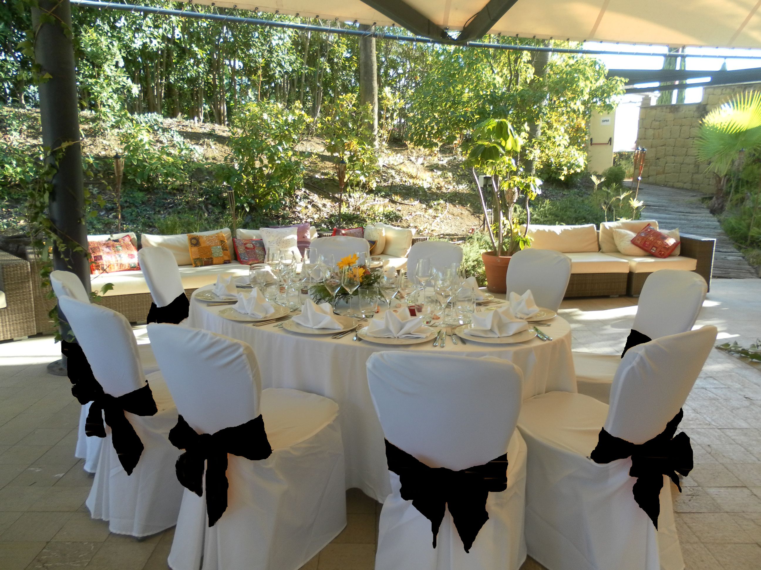 An outdoor dining area with round tables set for a meal, surrounded by chairs with black bows, and a sofa with cushions.