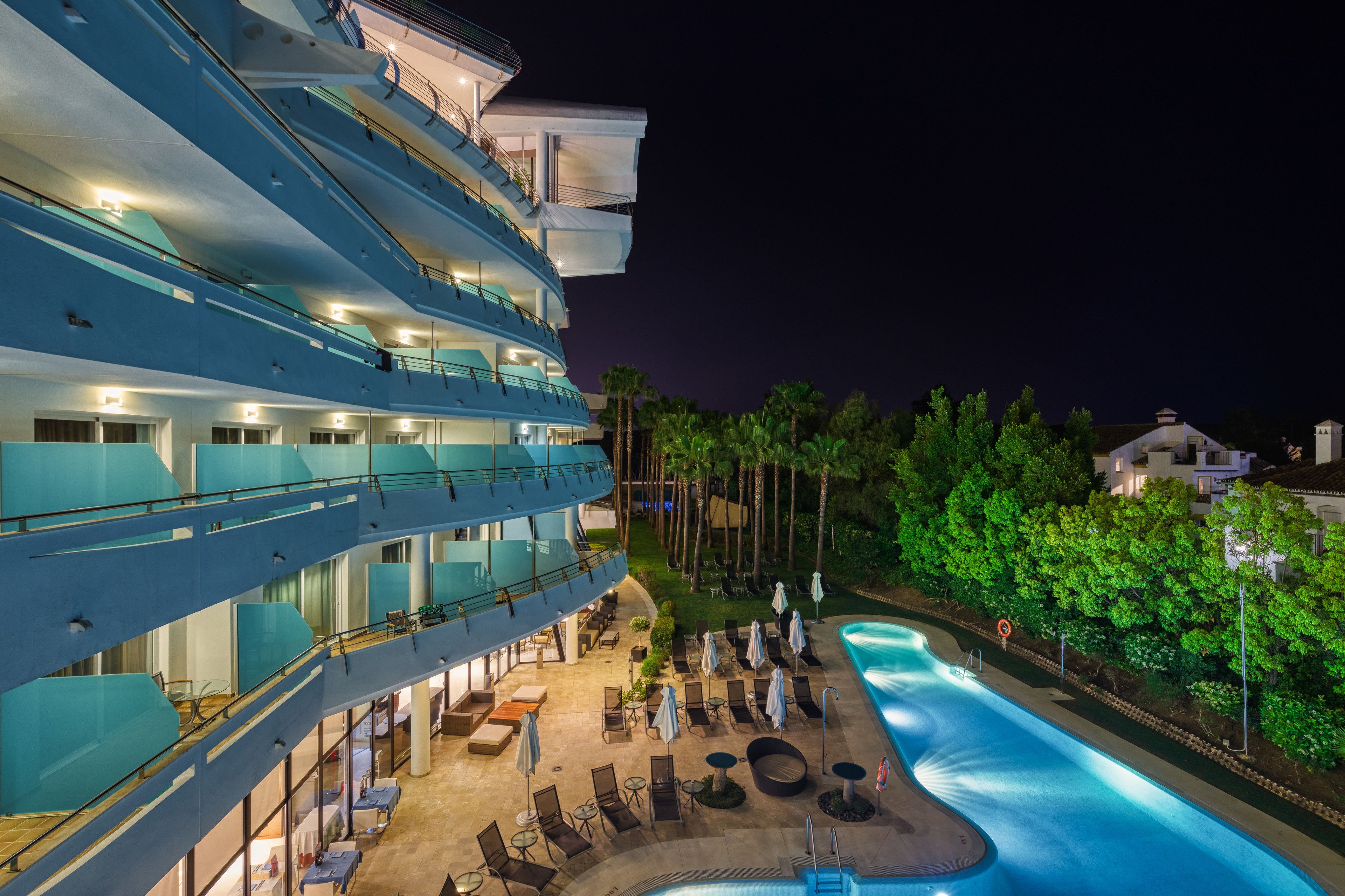 Un hotel de varios pisos con zona de piscina, mobiliario exterior y un balcón bien iluminado.