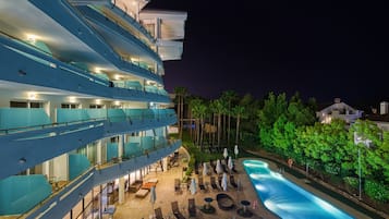 Un hotel de varios pisos con zona de piscina, mobiliario exterior y un balcón bien iluminado.