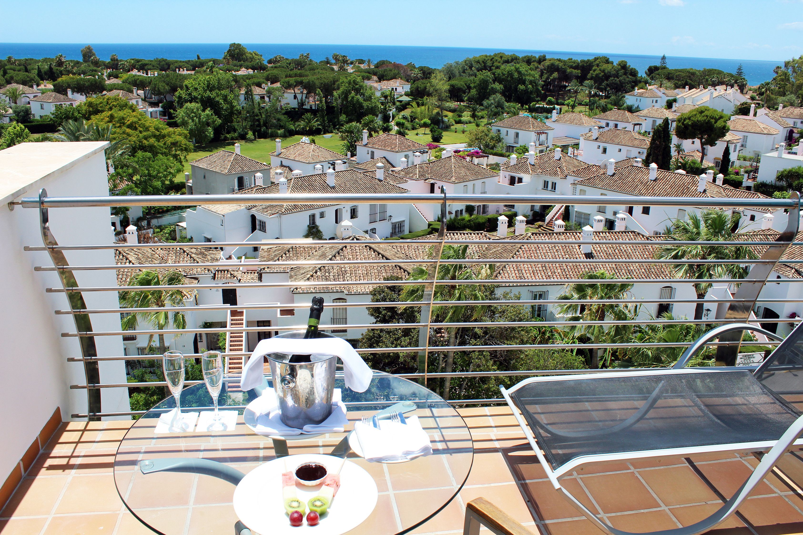 Un balcón con una mesa para dos, con vista a una zona residencial con casas y al mar.