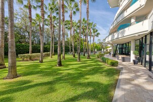 Un jardín bien cuidado con altas palmeras y un camino empedrado que lleva a un edificio moderno con amplios ventanales.