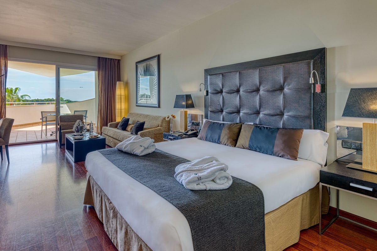 A hotel room with a large bed, a desk, a sofa, and a balcony with a view.