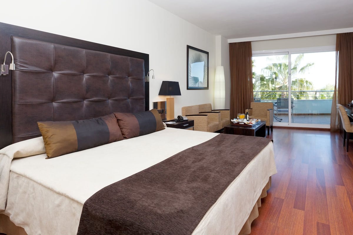 A hotel room with a large bed, a desk, a chair, and a balcony with a view of palm trees.