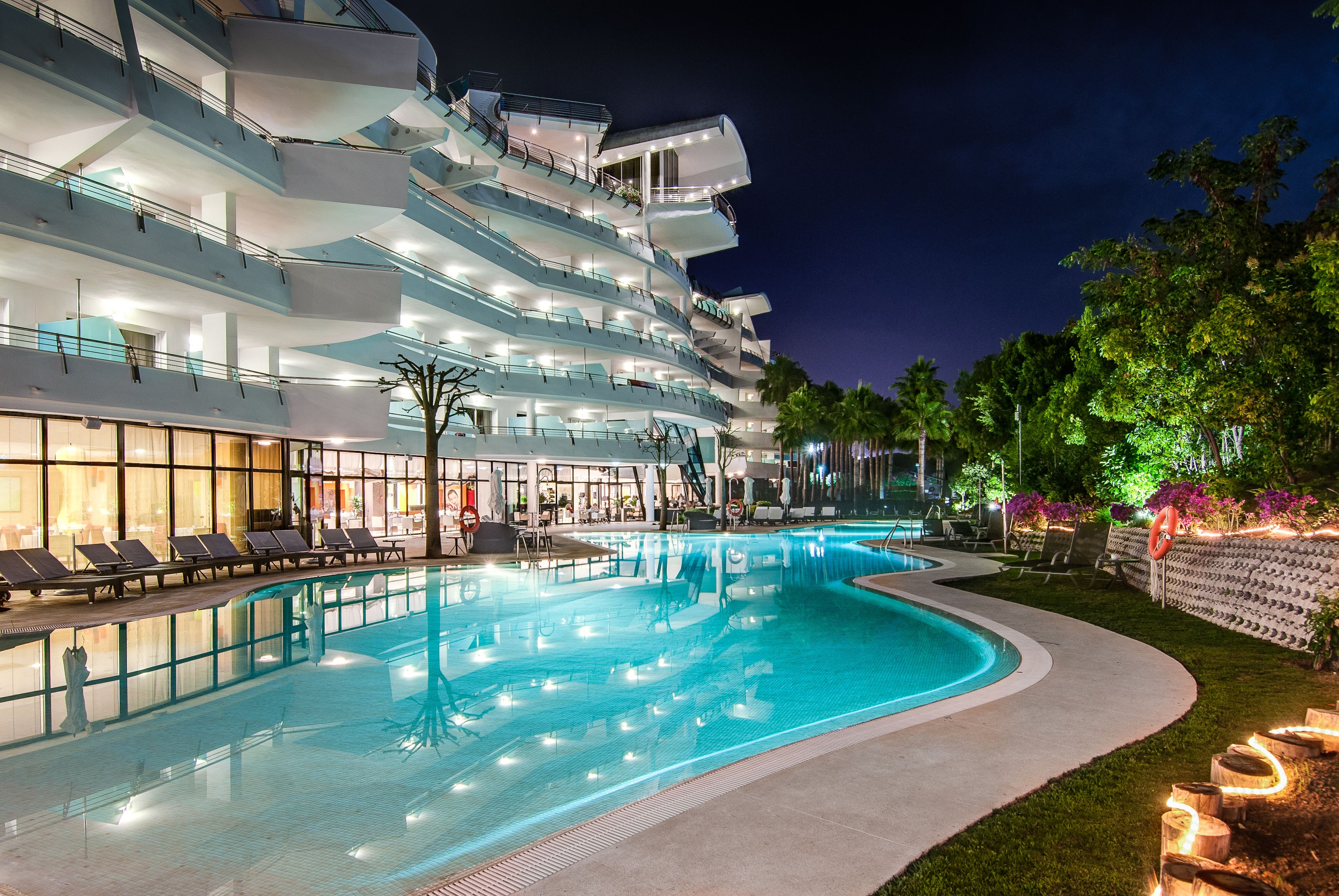 Ett modernt hotell med en swimmingpool, flera balkonger och ett väl upplyst utomhusområde.