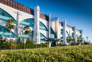 Een modern hotel met een gebogen glazen gevel, palmbomen en een helderblauwe lucht.