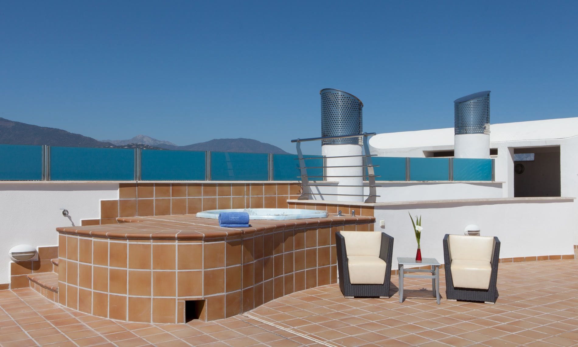 A rooftop terrace with a hot tub, two chairs, a table, and a view of the sea and mountains.