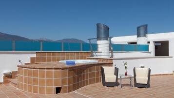 A rooftop terrace with a hot tub, two chairs, a table, and a view of the sea and mountains.