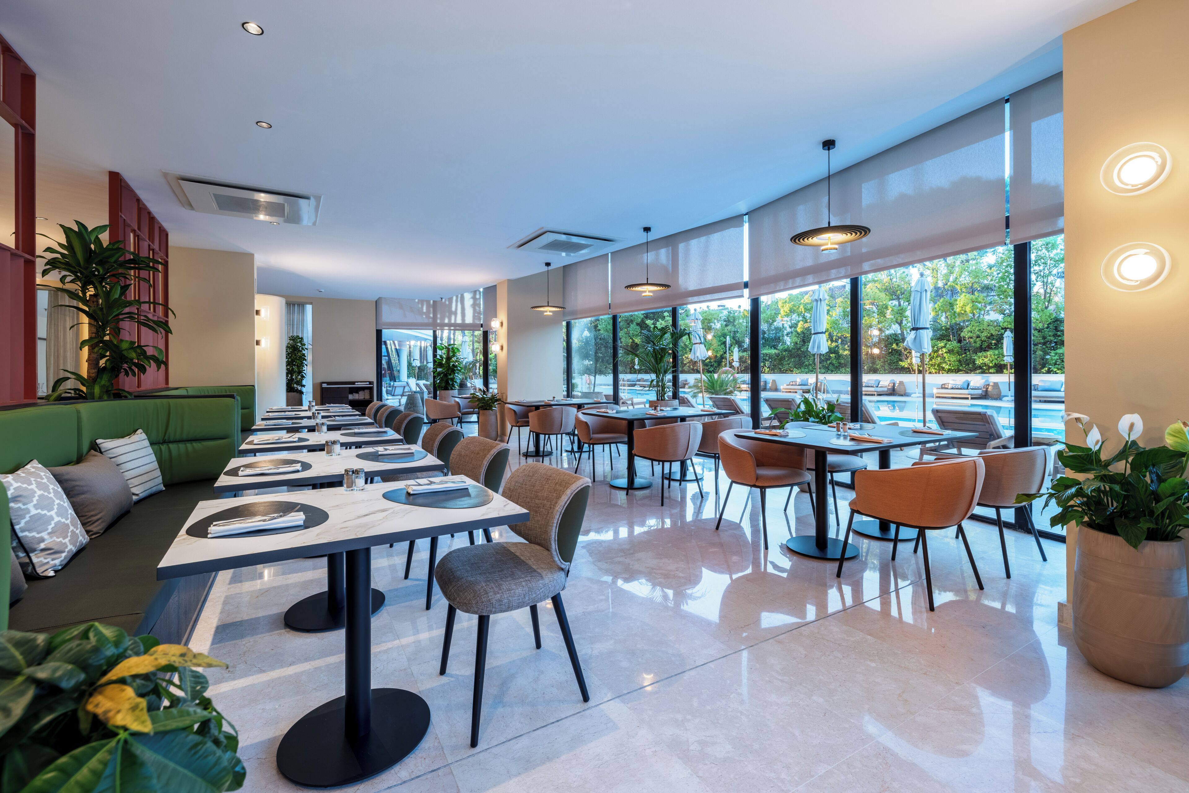Een modern restaurant met een lange toonbank, meerdere tafels en stoelen. Er staan potplanten en er is een groene zithoek.