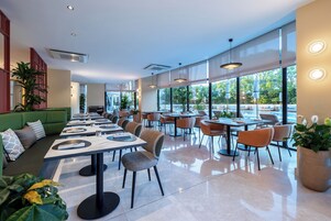 Een modern restaurant met een lange toonbank, meerdere tafels en stoelen. Er staan potplanten en er is een groene zithoek.