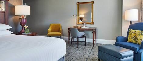 Een hotelkamer met een groot bed, een bureau met stoel, een blauwe fauteuil en een passende voetenbank.