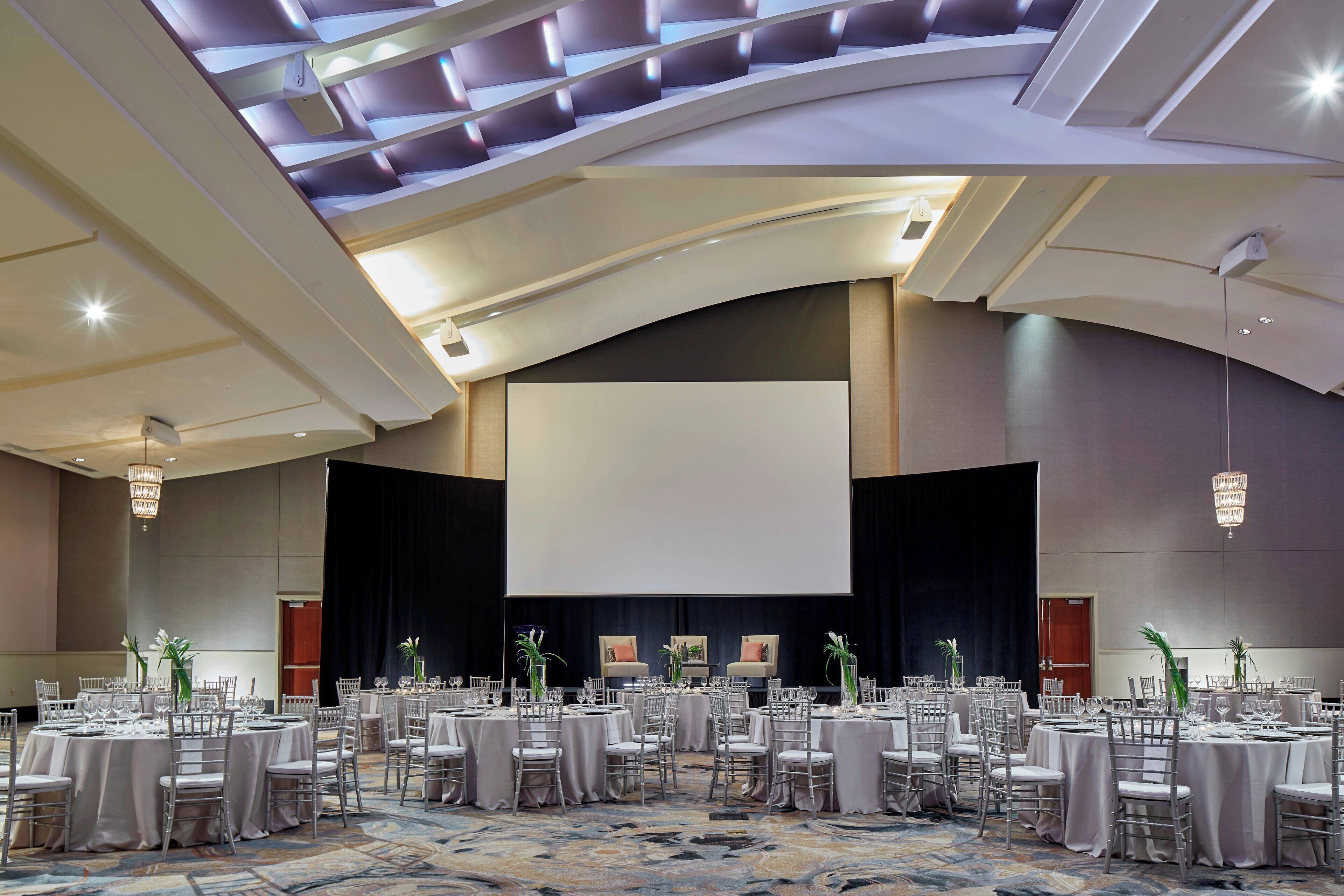 A large conference room with multiple round tables, chairs, and a large screen at the front.