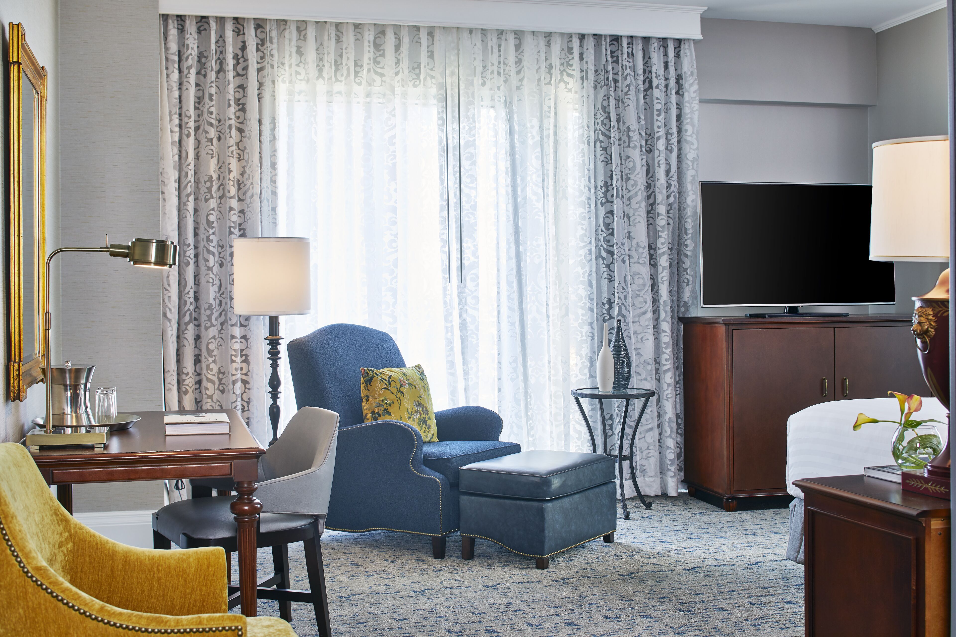 A hotel room with a large flat-screen TV, a blue armchair, a yellow armchair, a wooden desk, and a small side table.