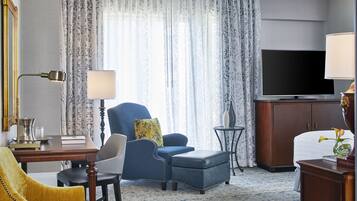 A hotel room with a large flat-screen TV, a blue armchair, a yellow armchair, a wooden desk, and a small side table.